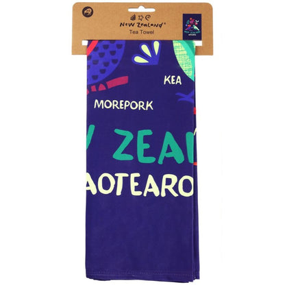 A New Zealand tea towel in its original cardboard packaging, featuring a deep purple background with colorful illustrations and the words 'KEA MOREPORK NEW ZEALAND MATEARO' in white, teal, and green.